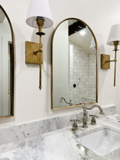 Gold or Brass Mirror between two brass sconces with shades. Over marble countertop and polished brass faucet.